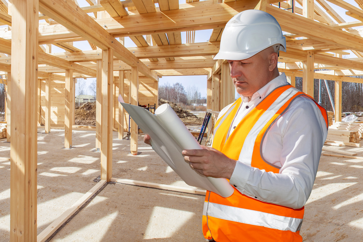 Architect wooden buildings. Man stands under frame wooden house. Man architect studies documentation or blueprints. Guy in protective helmet at construction site. Architect under timber framed house